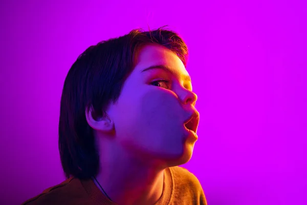 Detail malý chlapec, roztomilé dítě opírající se o průhledné sklo na tváři izolované na fialovém pozadí studia v neonovém světle — Stock fotografie