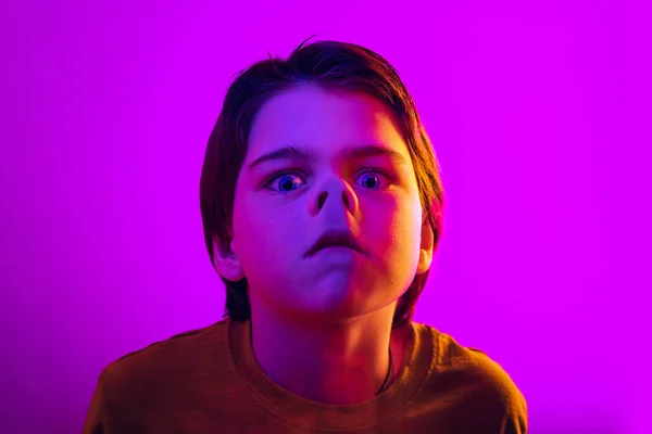 Close-up boy, funny child leaning against transparent glass by nose isolated on purple studio background in neon light — Foto de Stock