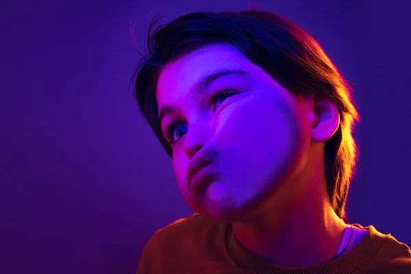 Close-up little boy, cute kid leaning against transparent glass by cheek isolated on purple studio background in neon light — Foto de Stock