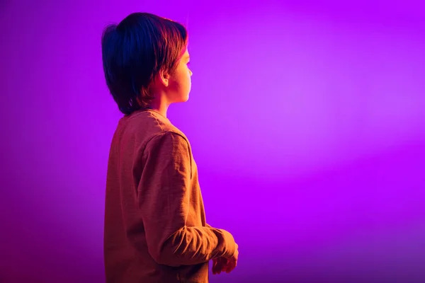 Side view portrait of boy, child dreamily looking away isolated over purple background in neon light — Stock Photo, Image