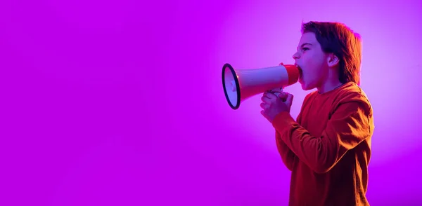 Portrait of boy, child shouting in megaphone, posing isolated over purple background in neon light. Flyer — Stock Photo, Image