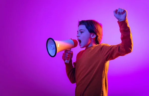 Portrait of boy, child shouting in megaphone, posing isolated over purple background in neon light — стоковое фото