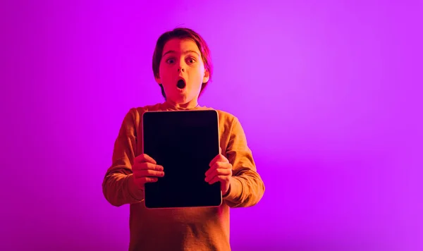 Retrato de menino, criança com emissões expressivas segurando tablet isolado sobre fundo rosa em luz de néon — Fotografia de Stock