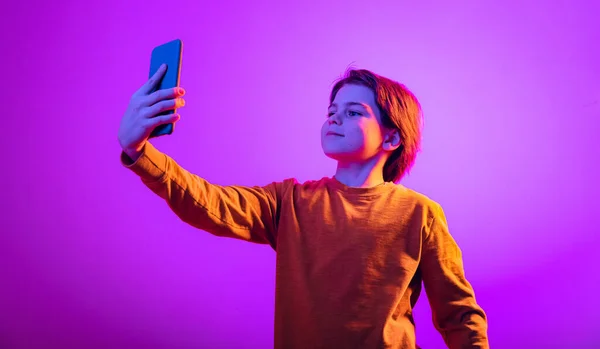 Portrait of boy, child taking selfie with phone isolated over pink background in neon light — Stock Photo, Image