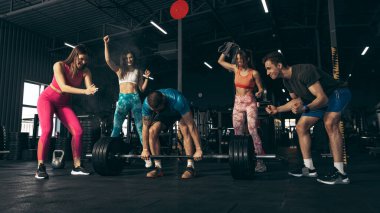 Spor yapan bir grup genç arkadaş, spor salonunda antrenman yapıyorlar. Ağırhalterleri kaldırmak için neşeli adam.