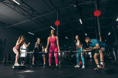 Spor yapan bir grup genç arkadaş, spor salonunda antrenman yapıyorlar. Spor kadını halter kaldırıyor, kabarık kasları var.