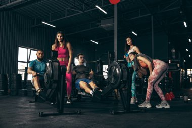 Spor yapan bir grup genç arkadaş, spor salonunda antrenman yapıyorlar. Çocuklar kürek çekme makinesinde egzersiz yapıyor. Eller antrenman günü