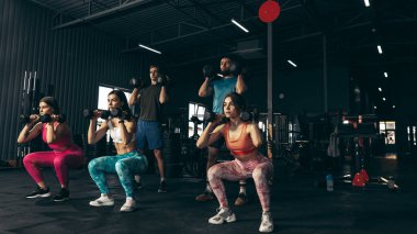 Spor yapan bir grup genç arkadaş, spor salonunda antrenman yapıyorlar. Sporcular dambılları kaldırıyor