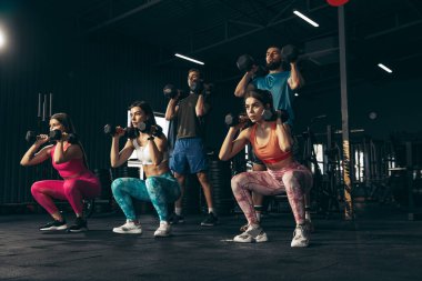 Spor yapan bir grup genç arkadaş, spor salonunda antrenman yapıyorlar. Sporcular dambılları kaldırıyor