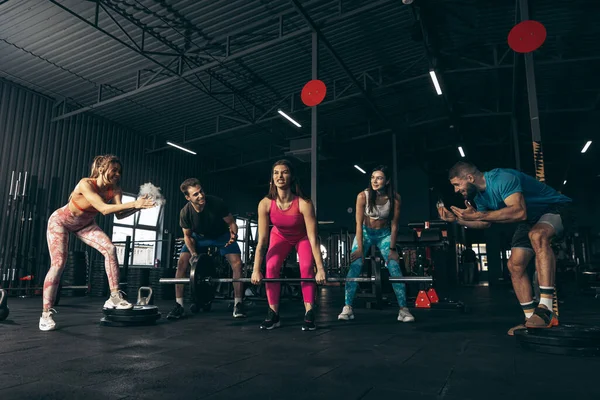 Grupo de jovens amigos fazendo esportes, treinando no ginásio dentro de casa. Mulher a levantar a campainha. Massa para cima músculos — Fotografia de Stock