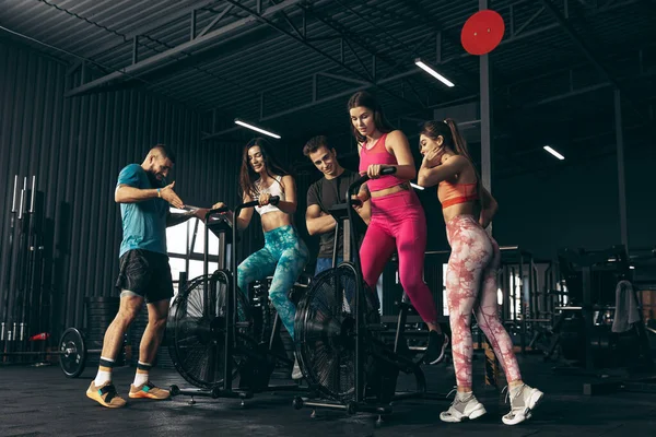 Grupo de jovens amigos fazendo esportes, treinando no ginásio dentro de casa. Treino de mulher em bicicleta de exercício. Dia de treinamento de perna — Fotografia de Stock