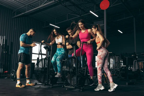 Grupper av unga vänner som idrottar, tränar på gym inomhus. Kvinnlig träning på motionscykel — Stockfoto