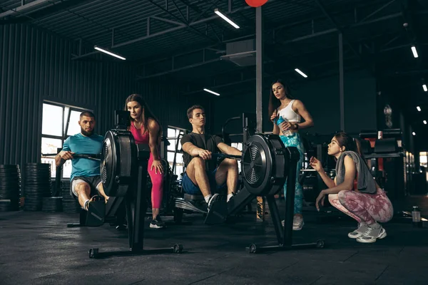 Spor yapan bir grup genç arkadaş, spor salonunda antrenman yapıyorlar. Erkekler kürek çekme makinesinde egzersiz yapıyor.