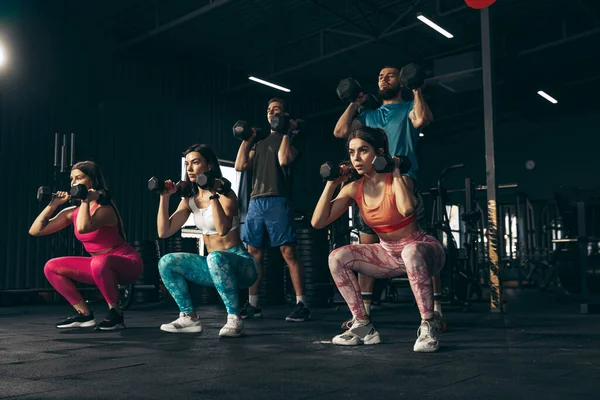 Spor yapan bir grup genç arkadaş, spor salonunda antrenman yapıyorlar. Sporcular dambılları kaldırıyor