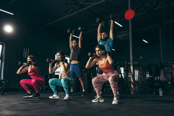 Spor yapan bir grup genç arkadaş, spor salonunda antrenman yapıyorlar. Sporcular dambılları kaldırıyor. Güçlü vücut