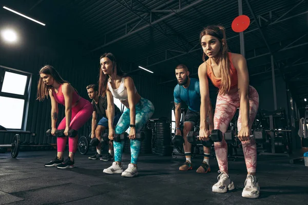 Grupper av unga vänner som idrottar, tränar på gym inomhus. Rygg- och handövningar med hantlar — Stockfoto