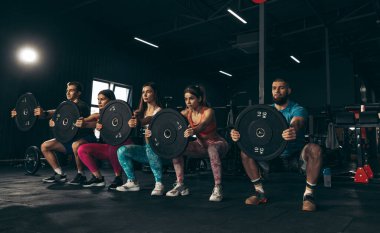 Spor yapan bir grup genç arkadaş, spor salonunda antrenman yapıyorlar. Ağırlıklarla çömelmek