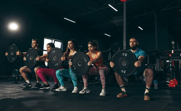 Spor yapan bir grup genç arkadaş, spor salonunda antrenman yapıyorlar. Ağırlıklarla çömelmek