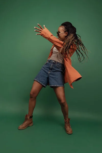 Menina africana emocional em shorts e dança camisa, se divertindo isolado em fundo verde escuro. Conceito de beleza, arte, moda, juventude e emoções — Fotografia de Stock