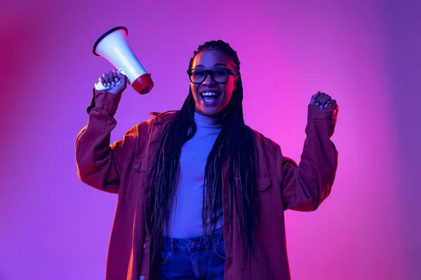 Happy african girl wearing warm shirt holding megaphone isolated on purple background in neon. Concept of emotions, beauty, art, fashion, youth, style — Fotografia de Stock