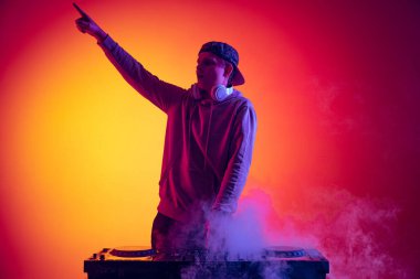 Portrait of active young man, professional dj playing, making sounds isolated over gradient red yellow background in neon light