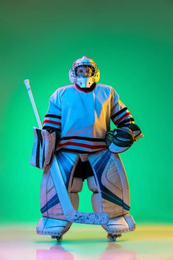 Genç bir çocuğun portresi, hokey oyuncusu özel koruyucu üniformalı kaleci yeşil arka planda izole edilmiş bir şekilde neon ışıkta poz veriyor..