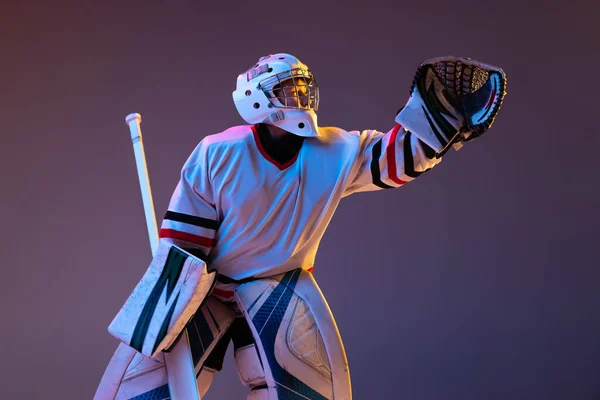 Portrait of teen boy, hockey player, goalkeeper catching plug in motion isolated over purple background in neon light. Dynamic sport — стоковое фото