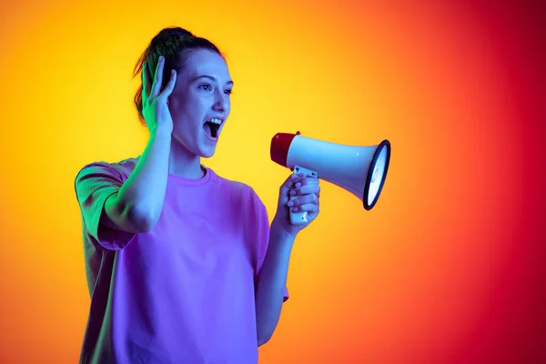 Beautiful excited girl shouting at megaphone isolated on yellow-red color background in neon light, filter. Concept of emotions, music, news, info — Stock Photo, Image