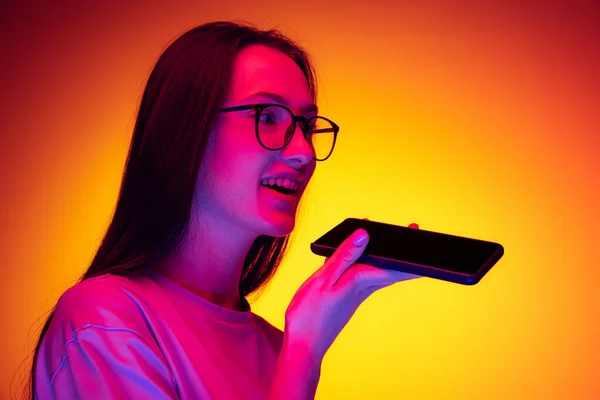 Portrait of young beautiful girl with long hair using phone isolated on orange background in neon light, filter. Concept of emotions, youth — Photo