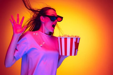 Heyecanlı bir genç kız, 3D gözlük takan ve elinde turuncu arka planda izole edilmiş patlamış mısır kovası tutan bir öğrenci neon ışığı, filtre. Duygular, gençlik, güzellik kavramı