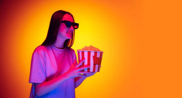 Young charming girl, student wearing 3d glasses and holding bucket of popcorn isolated on orange background in neon light, filter. Concept of emotions, youth, beauty — Stockfoto