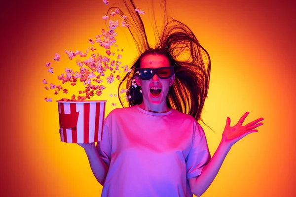 One excited young girl, student wearing 3d glasses and holding bucket of popcorn isolated on orange background in neon light, filter. Concept of emotions, youth, beauty — Stock fotografie