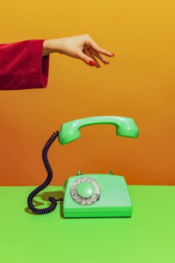 Colorful bright image of female hand holding old-fashioned green colored phone, handset falling down isolated over orange background