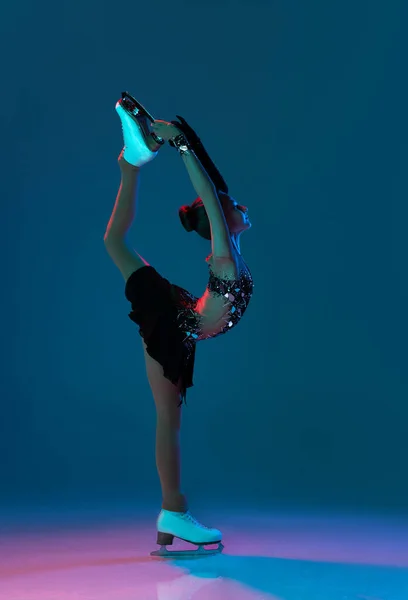 Portrait dynamique de jeune fille, patineuse artistique en robe de scène noire patinant isolée sur fond bleu au néon. Concept de sport, beauté, mode de vie actif. — Photo