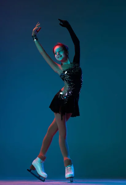 Dynamic portrait of young girl, female figure skater in black stage dress skating isolated on blue background in neon light. Concept of sport, beauty, active lifestyle. — Foto Stock