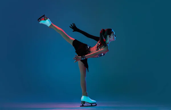 Portrait dynamique de jeune fille, patineuse artistique en robe de scène noire patinant isolée sur fond bleu au néon. Concept de sport, beauté, mode de vie actif. — Photo