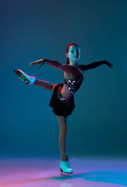 Dynamic portrait of young girl, female figure skater in black stage dress skating isolated on blue background in neon light. Concept of sport, beauty, active lifestyle. — Stock Photo, Image