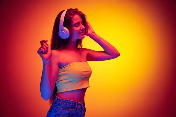 Half-length portrait of young girl listening to music in headphones isolated over gradient red yellow background in neon — Zdjęcie stockowe