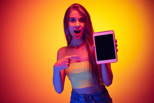 Портрет молодой красивой девушки, указывающей с шокированным выражением лица на планшете, выделенный над градиентным оранжевым желтым фоном в неоне — стоковое фото