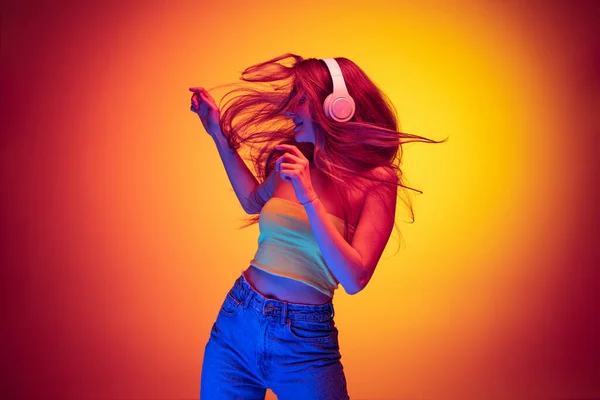 Портрет молодої красивої дівчини, що слухає музику в навушниках і танцює ізольовано на градієнтному червоно-жовтому фоні в неоні — стокове фото