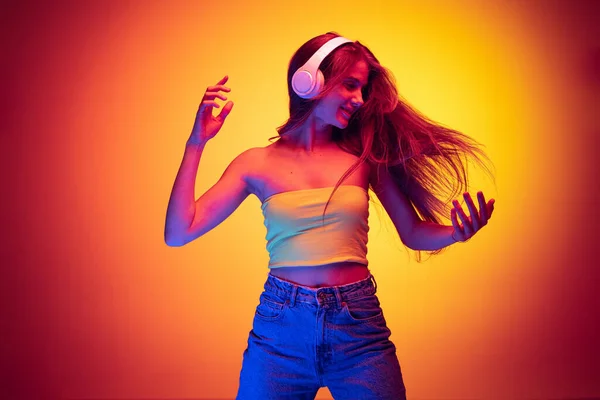Портрет молодої емоційної дівчини, що слухає музику в навушниках і танцює ізольовано на градієнтному червоно-жовтому фоні в неоні — стокове фото