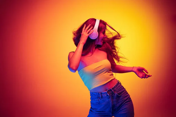 Portrét mladé šťastné dívky poslech hudby ve sluchátkách a tanec izolované přes gradient červené žluté pozadí v neonu — Stock fotografie