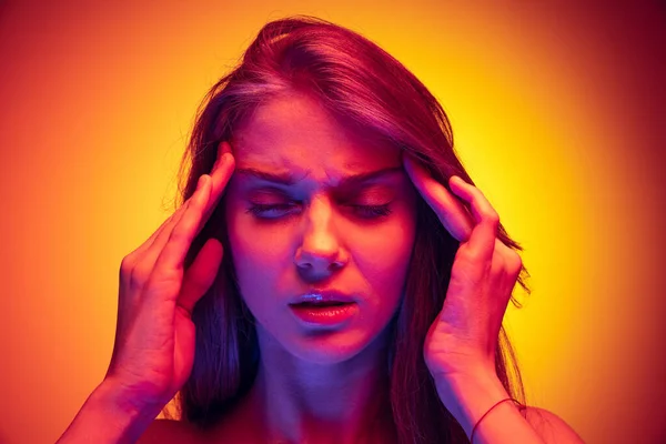 Close-up portrait of young girl touching temples, suffering from headache isolated over gradient red yellow background in neon — Stockfoto