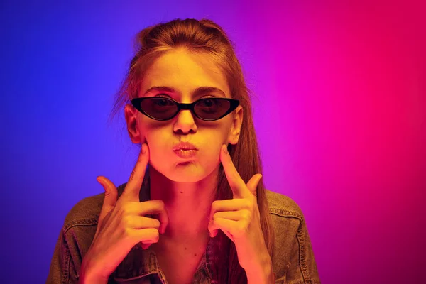Portrait of young woman in stylish sunglasses making funny face isolated over blue pink background in neon light — Stock fotografie
