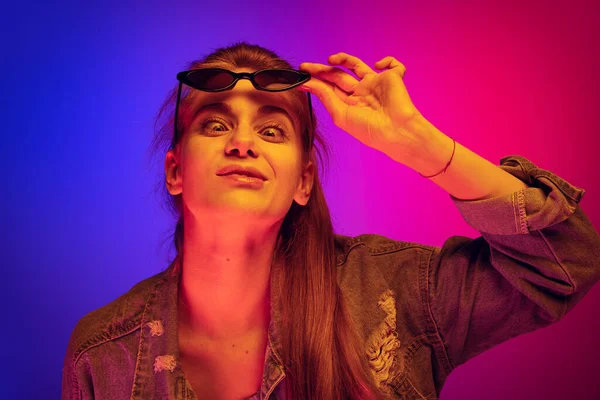 Портрет молодої дівчини, що піднімається сонцезахисними окулярами вгору і робить смішні обличчя ізольовані над градієнтним синьо-рожевим фоном в неоновому світлі — стокове фото