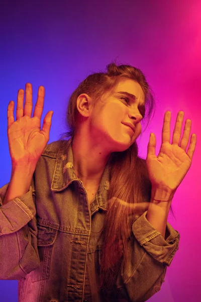 Grimace. Young funny girl leaning against transparent glass isolated over gradient blue pink background in neon light. — Stock Fotó