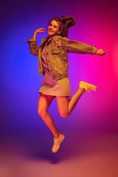 Portrait of young girl in casual cloth jumping, posing isolated over gradient pink blue background in neon light — Zdjęcie stockowe