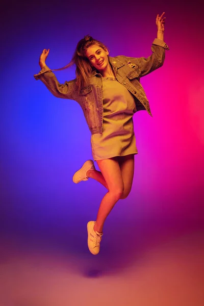 Portrait of young girl in casual cloth jumping, posing isolated over gradient pink blue background in neon light. Cheerful look — Zdjęcie stockowe