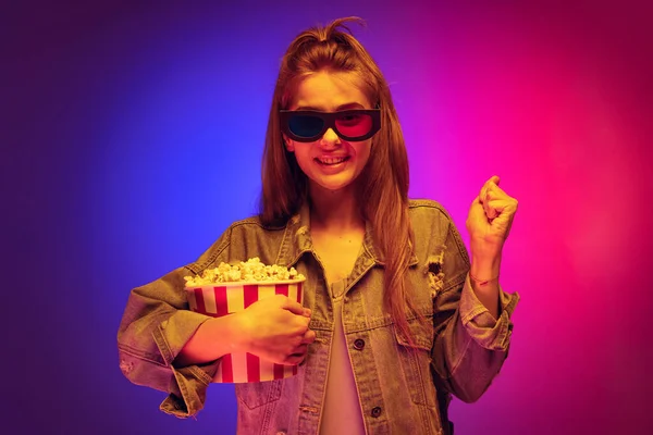 Портрет молодої дівчини в джинсовій куртці в окулярах 3D-кіно і тримає попкорн кошик ізольований на градієнтному рожево-фіолетовому фоні в неоні — стокове фото