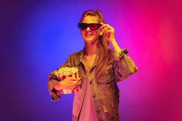 Portrait of young girl in denim jacket wearing 3D cinema glasses and holding popcorn basket isolated over gradient pink purple background in neon — Stock fotografie